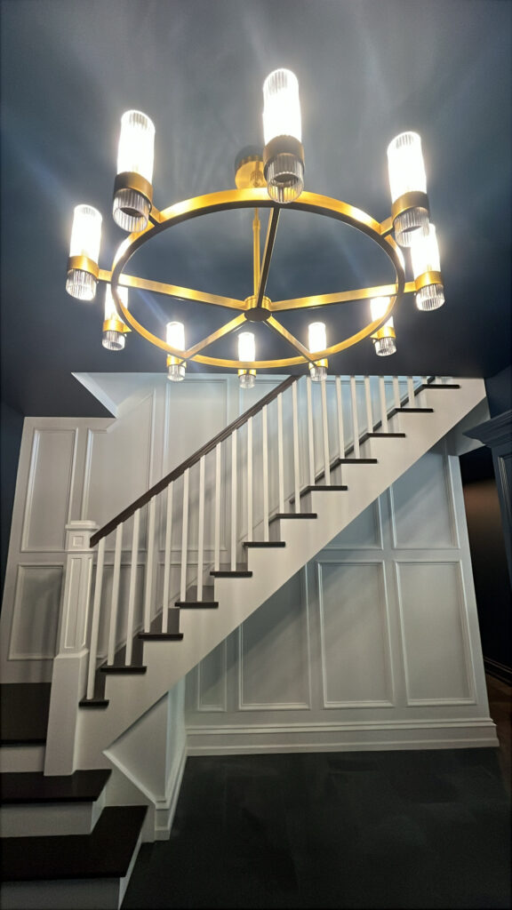 Stair hall in Scarsdale, NY with a dark ceiling, white paneling, and a dramatic chandelier above the staircase