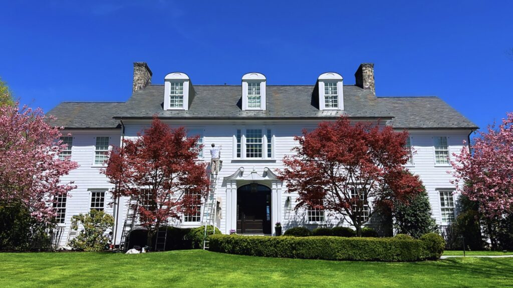 Classic white colonial home in Darien, CT with painters working on the front elevation, dark entry door, and vibrant spring landscaping